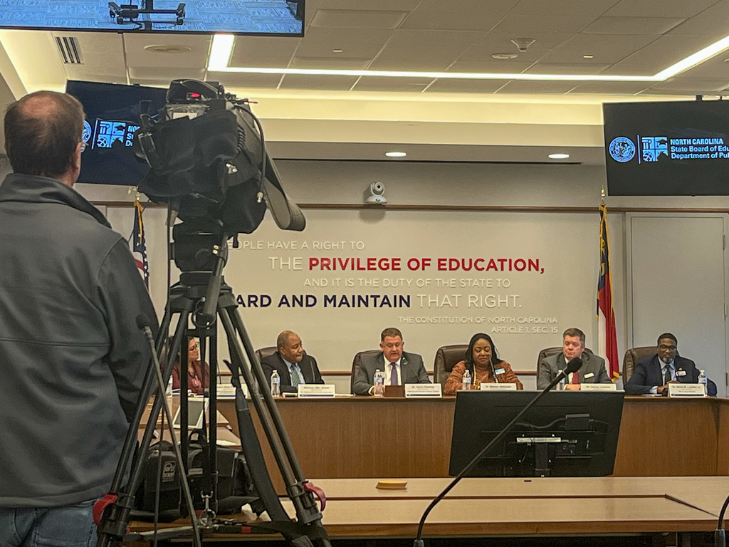 A camera operator films the North Carolina State Board of Education members seated at a panel table during a press conference, with the state motto "The privilege of education" displayed on the wall behind them.