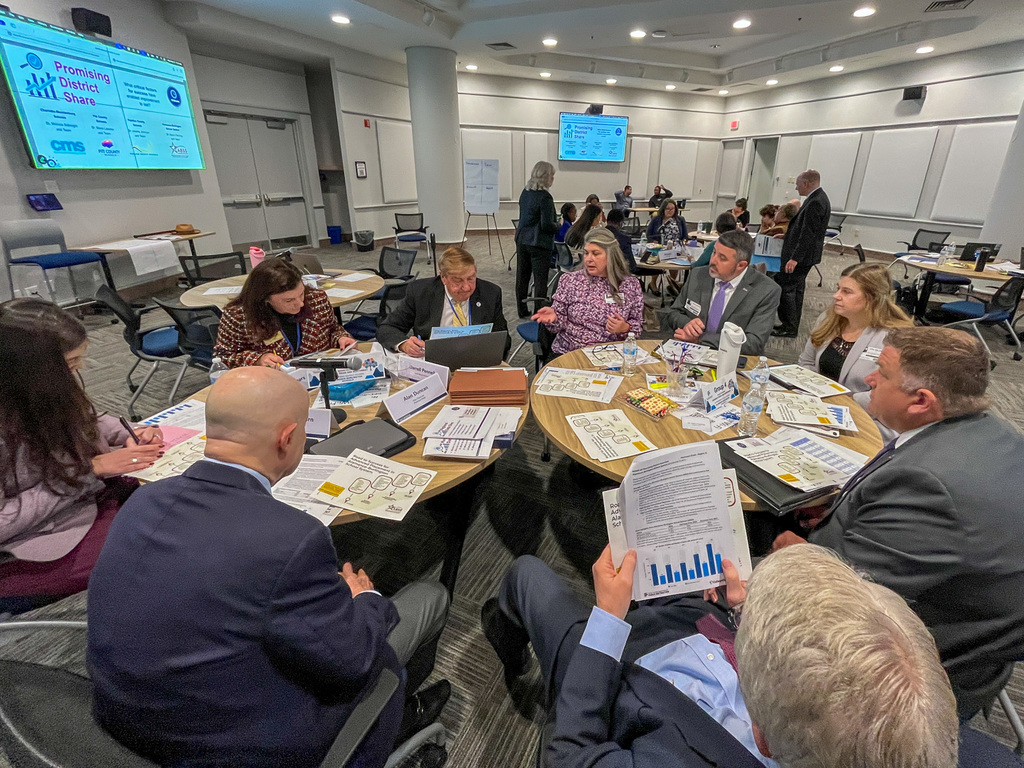 Education leaders from multiple school districts seated around tables in a collaborative work session, reviewing documents and data with presentations displayed on screens in the background at the State Board of Education meeting.