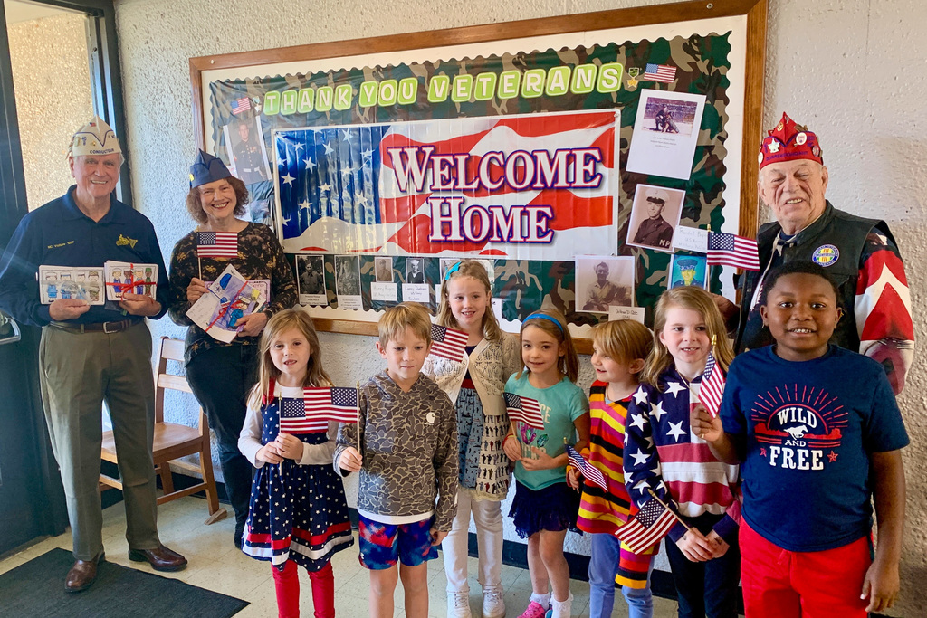 Veterans and first-grade students pose in front of a "Welcome Home" bulletin board honoring veterans and displaying photos of service members.