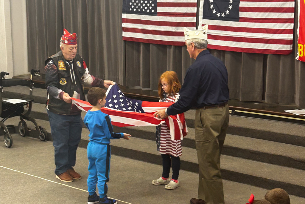 Two veterans demonstrate proper flag folding technique to young first-grade students during a Veterans Day program, with American flags displayed in the background.