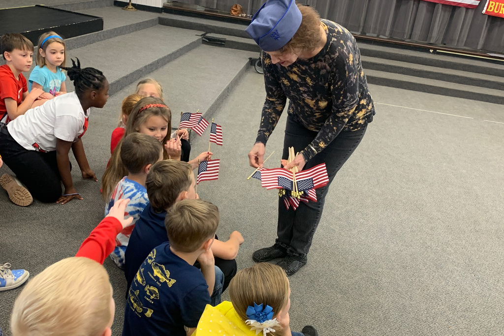A veteran distributes small American flags to excited first-grade students seated on the floor during the flag education program.