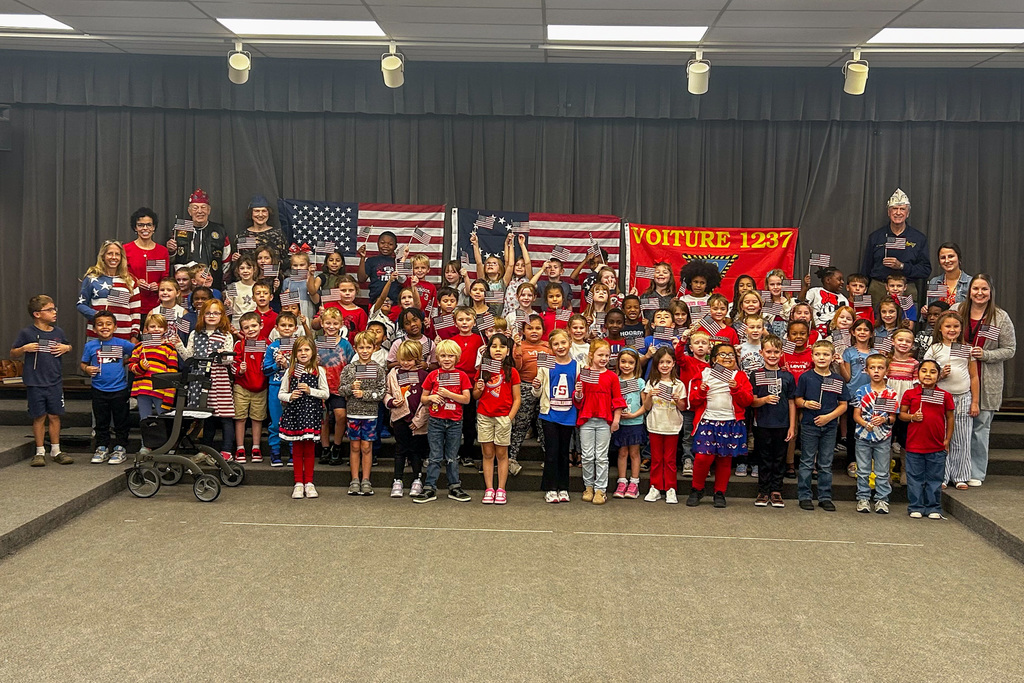 Large group photo of all E.M. Holt Elementary first graders, teachers, and veterans holding American flags with the Voiture 1237 banner displayed prominently.