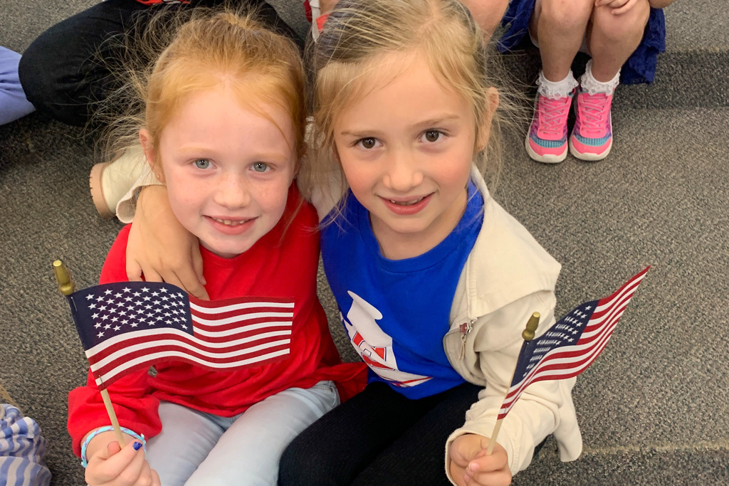 Two smiling first-grade girls sit together on the floor proudly holding their American flags.