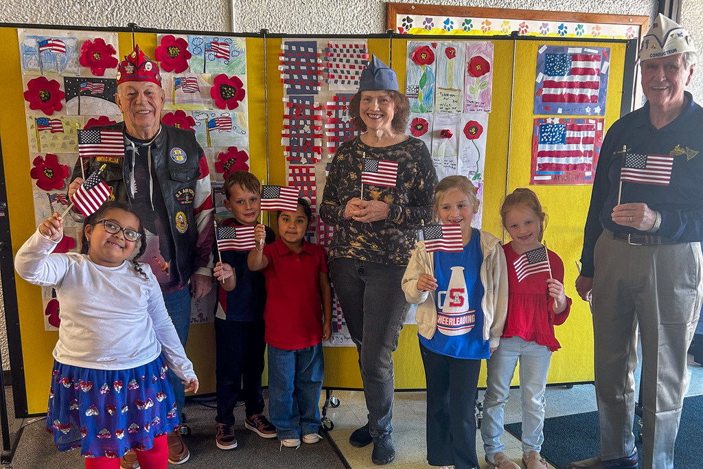 Veterans pose with enthusiastic first-grade students holding American flags in front of classroom bulletin boards featuring patriotic student art projects.