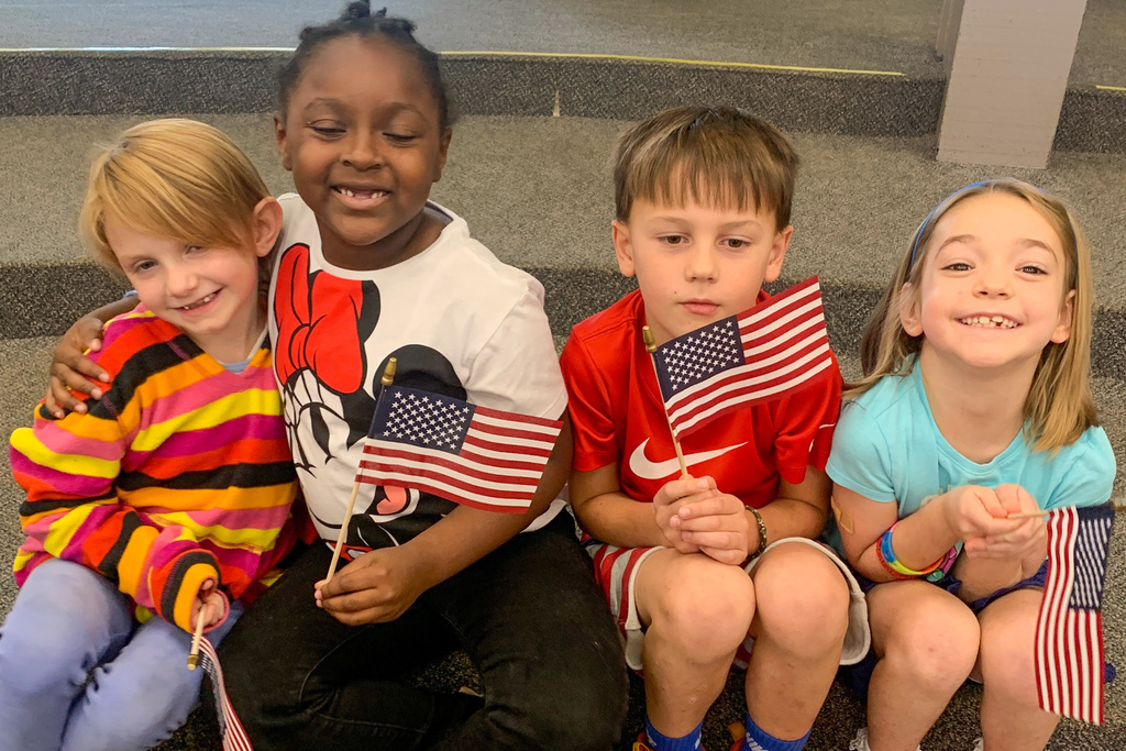 Four diverse first-grade students smile together while holding their American flags.