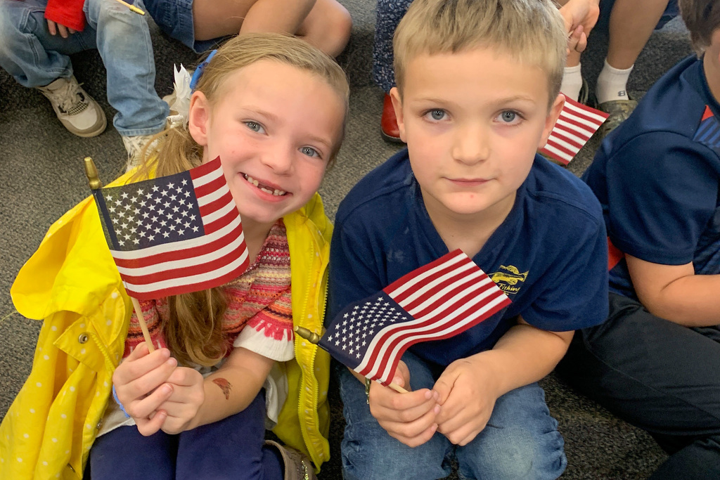 Two first-grade students seated on the floor display their new American flags during the Veterans Day program.