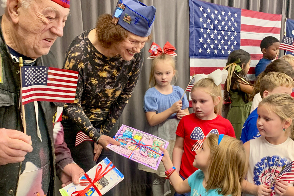 Veterans interact with first-grade students, showing them thank-you cards and distributing flags during the ceremony.