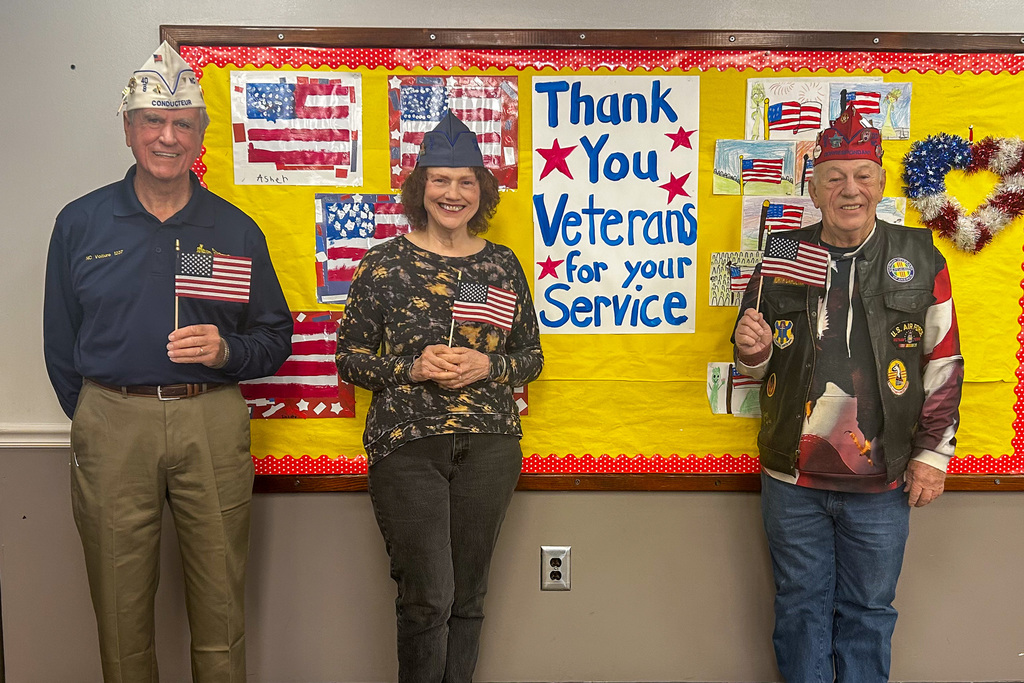 Three veterans stand together holding small American flags in front of a "Thank You Veterans for your Service" bulletin board decorated with student artwork.