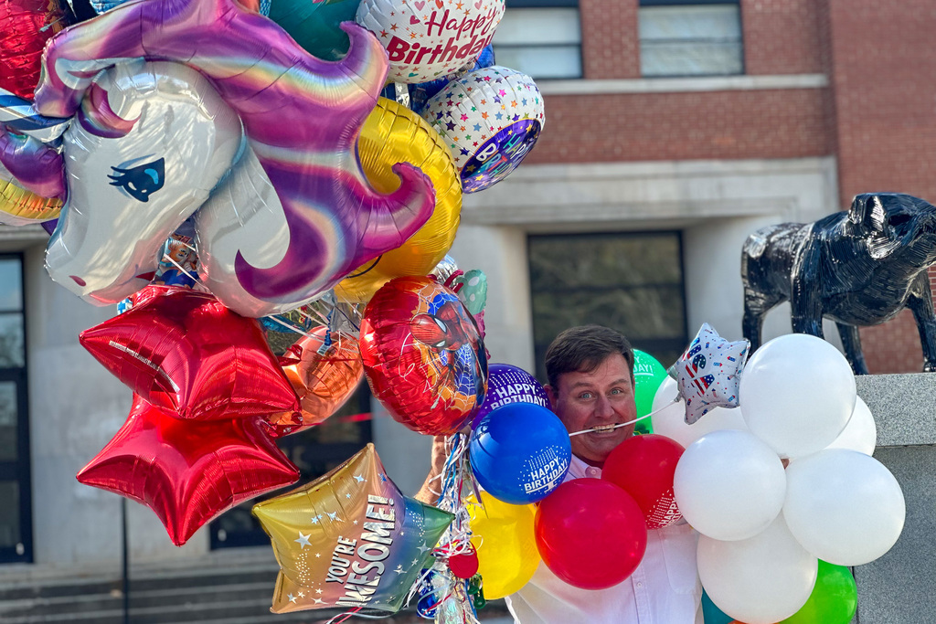 A close-up shot of the man (Principal Bryan) surrounded by a huge, overflowing cluster of foil and latex balloons, including a large unicorn-shaped balloon and several star-shaped balloons. He is peeking out from behind the balloons with a humorous, surprised expression.