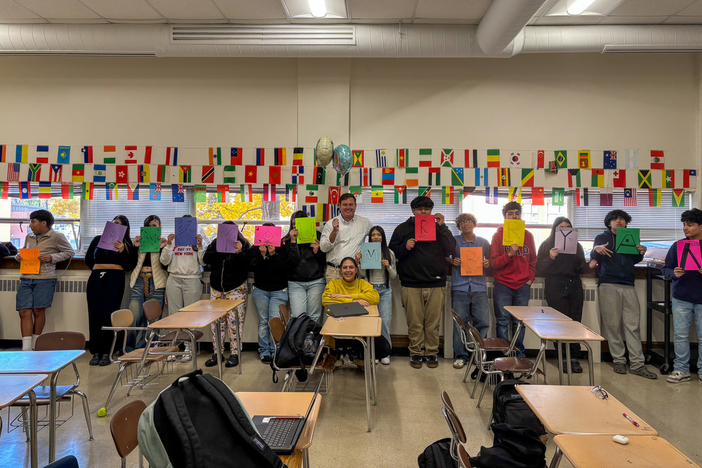 A group of high school students and two teachers standing in a classroom, with a colorful banner of international flags behind them. The students are holding up colored pieces of paper that, together, spell out the principal's name.