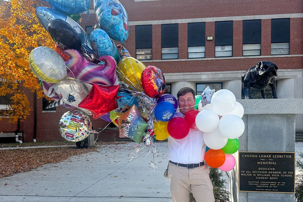 A man (Principal Bryan) standing outdoors holding a massive bouquet of brightly colored Mylar and latex balloons, including unicorn, stars, and cartoon-themed shapes. He is mostly obscured by the balloons and has a surprised expression. A brick building and a memorial statue are in the background.