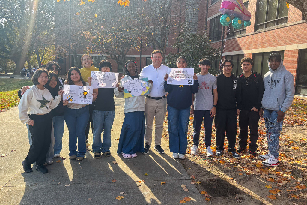 A group of high school students and two teachers posing outdoors on a sunny day. They are holding handmade signs with positive messages and paw print images. The school building is in the background, and autumn leaves cover the ground.