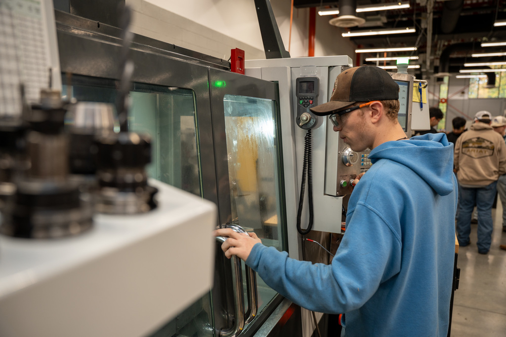A high school student in a blue hoodie and cap operates a commercial-grade piece of equipment with a glass door and control panel in a professional kitchen or food service training facility.