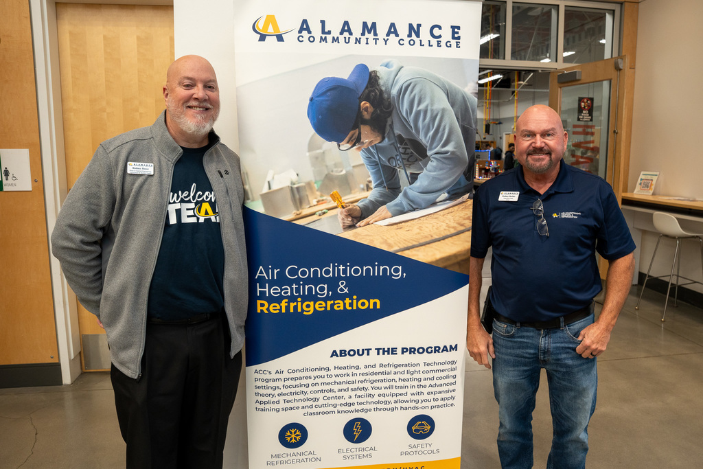 Two Alamance Community College staff members pose on either side of a promotional banner for the Air Conditioning, Heating, and Refrigeration program, which features an image of a student at work and lists program focus areas including mechanical refrigeration, electrical systems, and safety protocols.