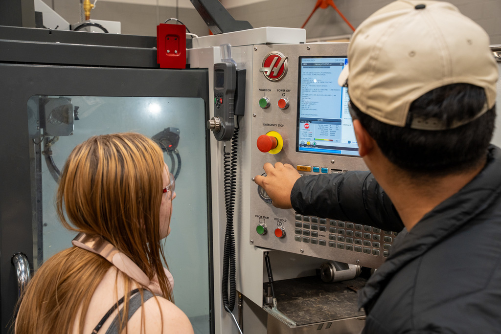 Two students lean over a CNC machine control panel, with one student pointing at the digital screen displaying programming code while an instructor observes their work.
