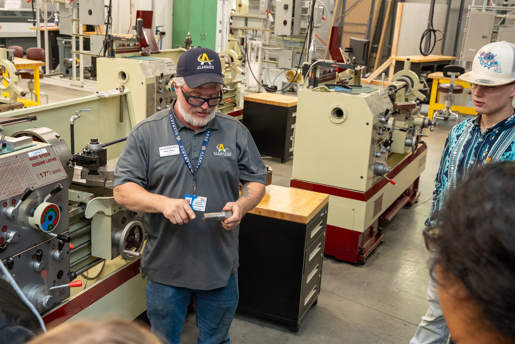 An Alamance Community College instructor wearing a branded cap and polo shirt demonstrates precision measurement techniques with calipers on a threaded metal part to a student in a machine shop filled with lathes and manufacturing equipment.
