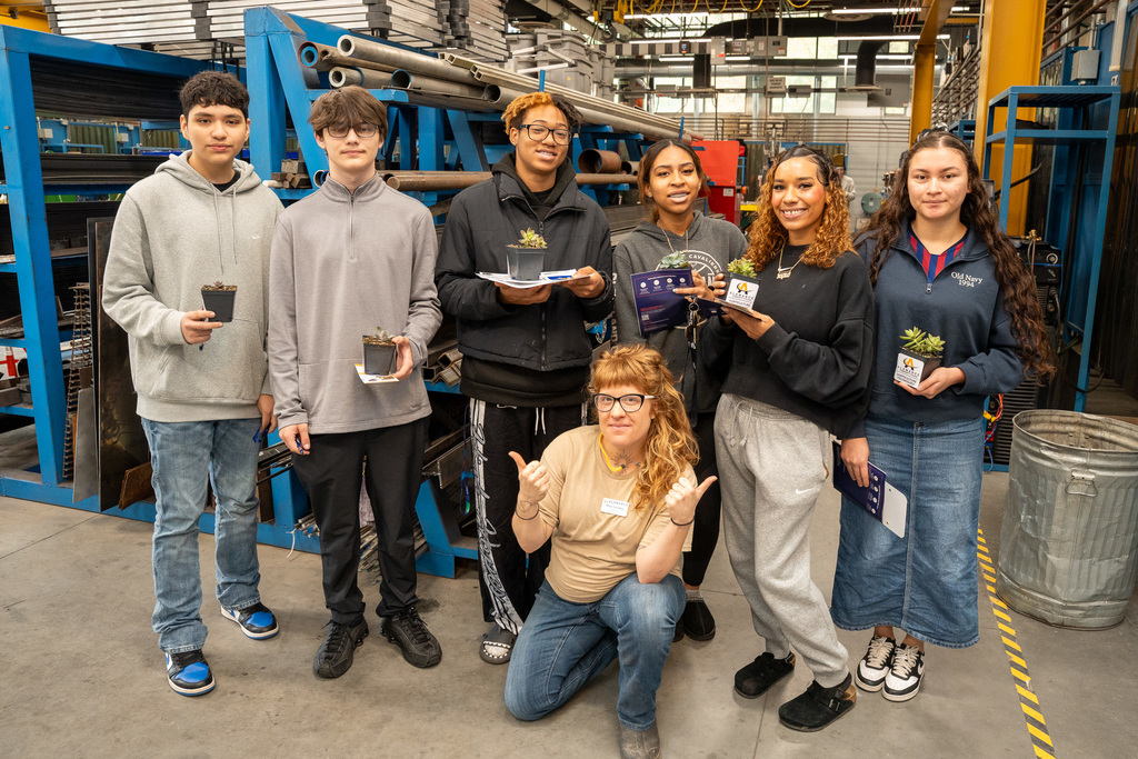 A diverse group of seven high school CTE students pose together in an industrial manufacturing workshop, holding small potted plants and informational materials, with blue shelving and metal storage racks visible behind them.