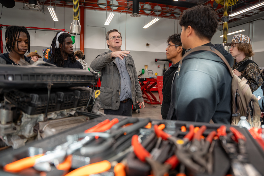 An instructor gestures while explaining automotive concepts to a group of attentive high school students gathered around engine components and tools in a well-equipped automotive technology lab.