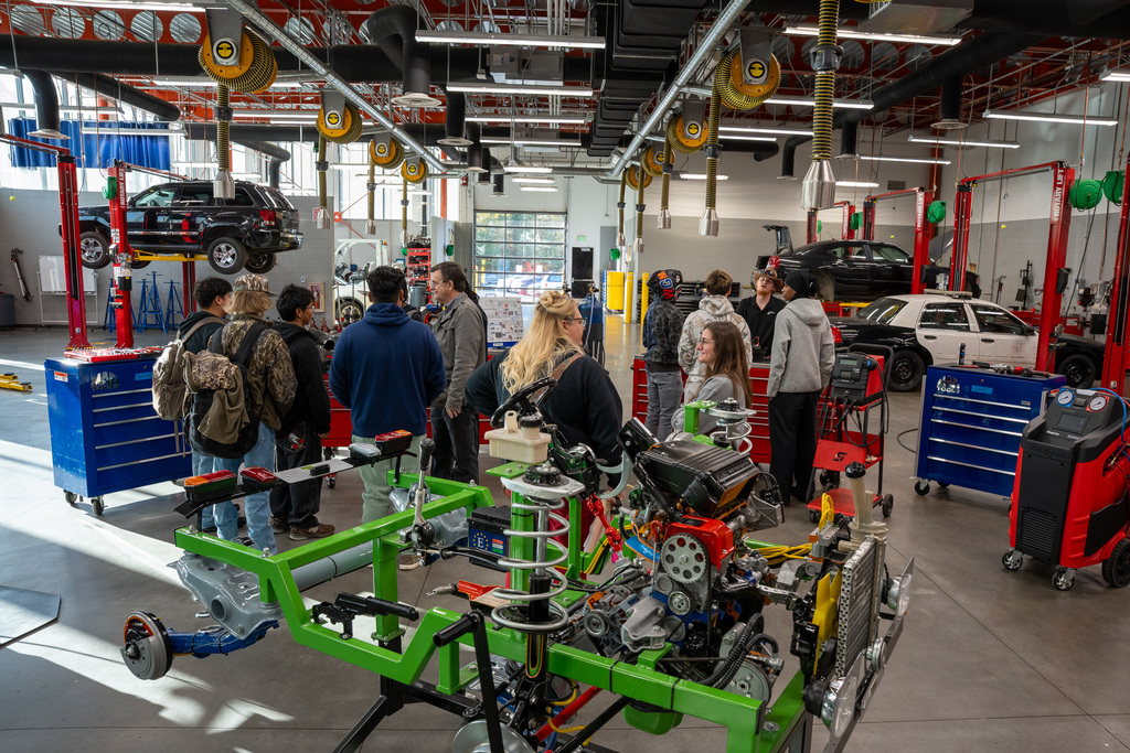 High school students gather in a modern automotive repair facility with vehicle lifts, examining a disassembled engine mounted on a bright green educational display stand, while cars are elevated on lifts in the background.