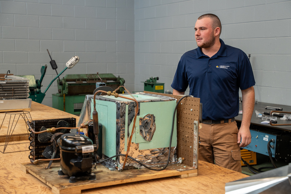 A male instructor in an Alamance Community College polo shirt stands in a workshop next to disassembled HVAC equipment including compressors and refrigeration units on a workbench, with industrial machinery visible in the background.