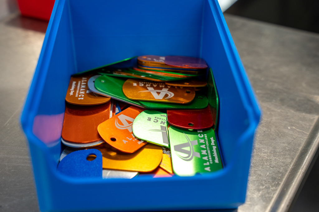 A blue plastic bin filled with colorful anodized aluminum keychains and tags featuring the Alamance Community College logo in various bright colors including orange, green, yellow, and purple.