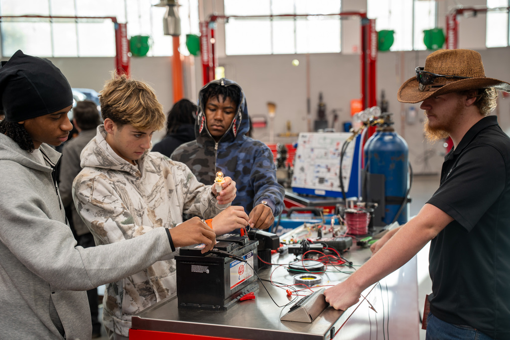 Students work hands-on with automotive batteries and electrical testing equipment on a workshop table, guided by an instructor in a cowboy hat, with welding tanks and automotive repair equipment visible in the background.