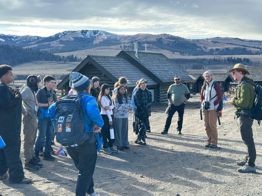 Park ranger or guide speaking to a group of middle school students outdoors at Yellowstone with historic buildings and mountains visible behind them