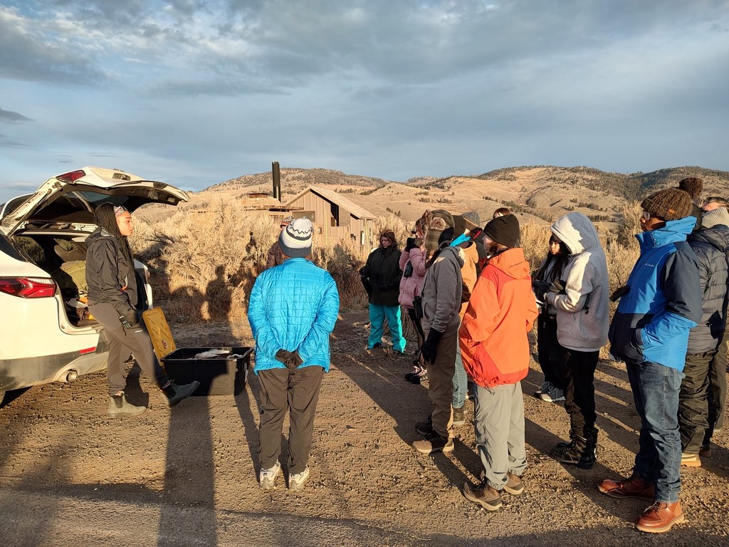 Students gathered in a circle outdoors listening to an instructor during a field lesson in the Yellowstone area with rolling hills in the background