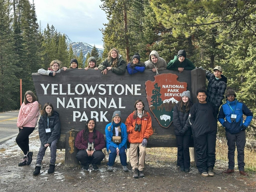 Group of middle school students and teacher posing at the Yellowstone National Park entrance sign with mountains and evergreen trees in the background
