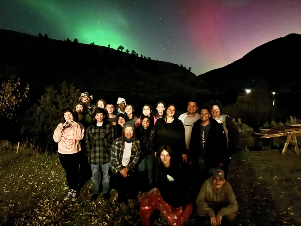 Group photo of students and teacher at night with the Northern Lights displaying green and pink colors in the sky above mountains