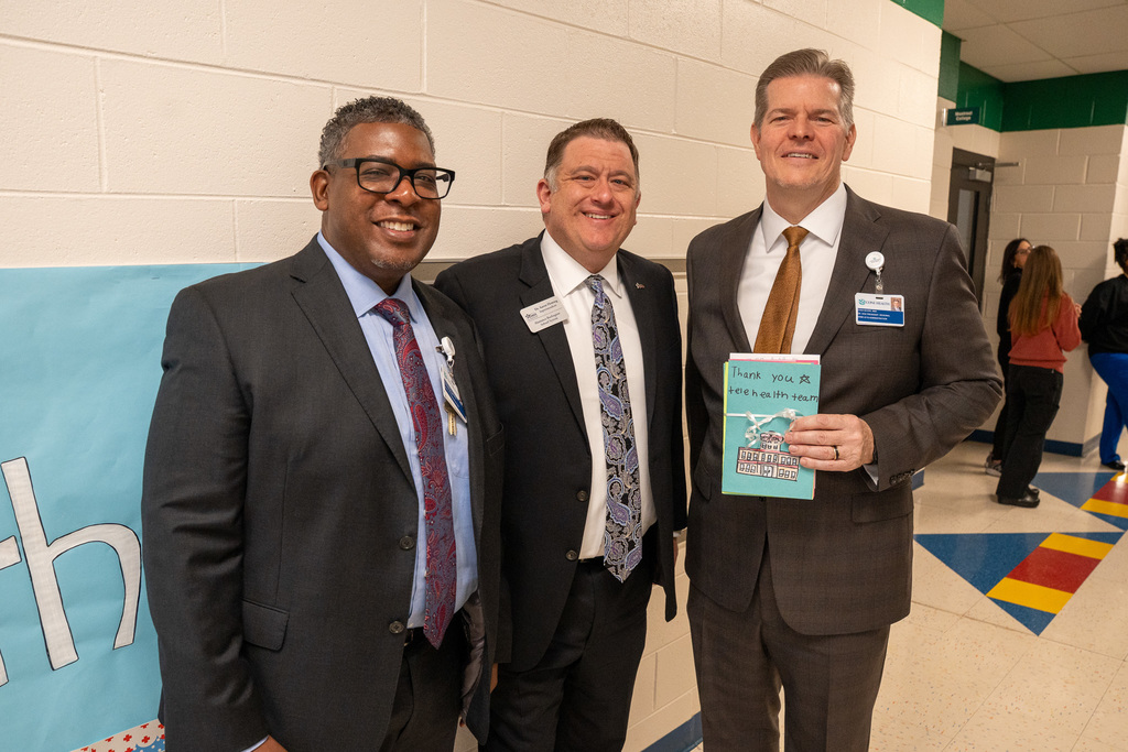 Three men in suits and ties standing together in a school hallway. The man on the right holds a small, colorful, handmade "Thank you to telehealth team" booklet. These are likely officials and partners celebrating the program launch.
