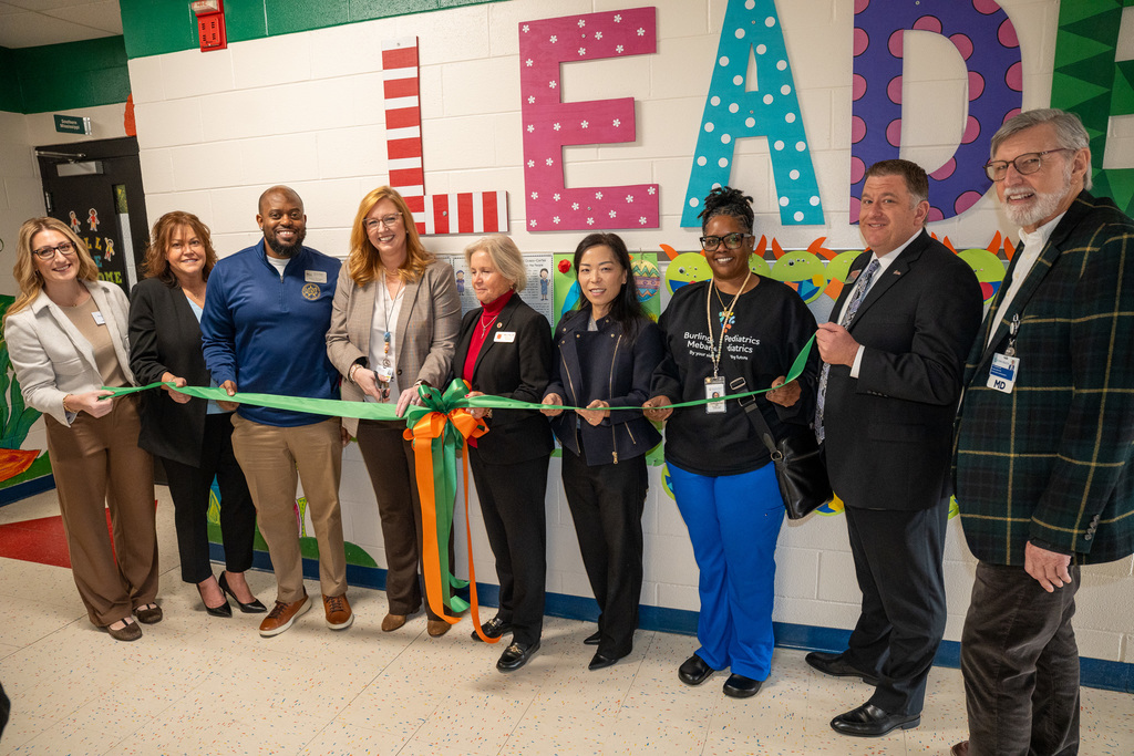 A group of nine individuals, including school officials, Cone Health representatives, and community partners (including Dr. Fleming and Principal Ray), posing for a ribbon-cutting ceremony in the school hallway to officially launch the telehealth program.