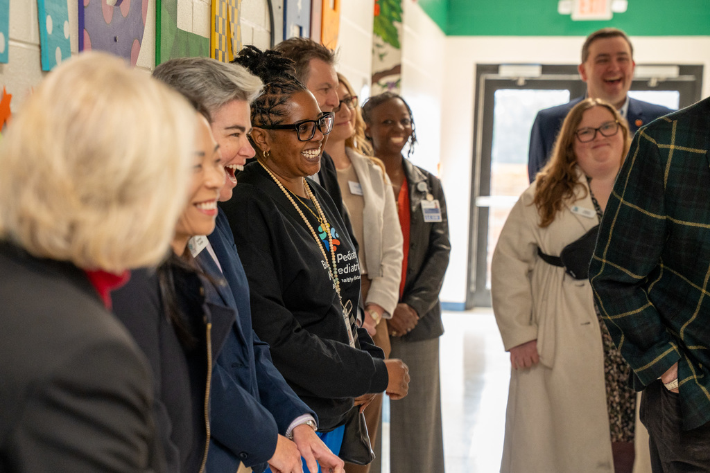 A candid, joyful group shot of several adults—including school and health officials—smiling and laughing while standing in a line in the school hallway.