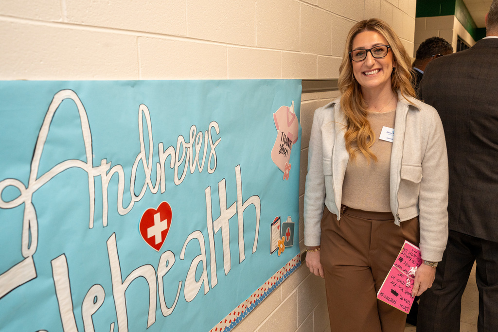 A woman in a light jacket and brown pants smiles while standing next to a large blue handmade sign on the wall that reads "Andrews Telehealth" with a red heart.