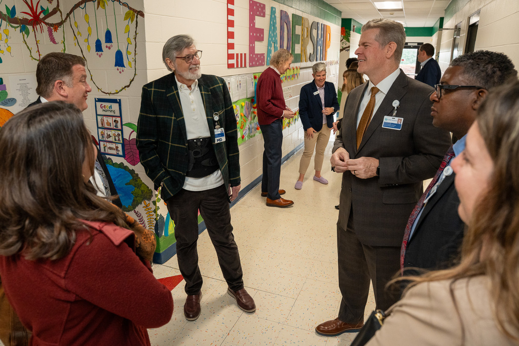 A group of officials, including a man in a distinguished plaid jacket, standing and engaging in a conversation in the school hallway during the telehealth program launch event.