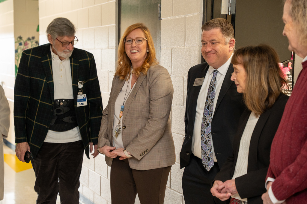 Three adults—a man in a plaid jacket, a smiling woman in a tan blazer, and a man in a suit—standing and talking in a small group near a door in the school hallway.