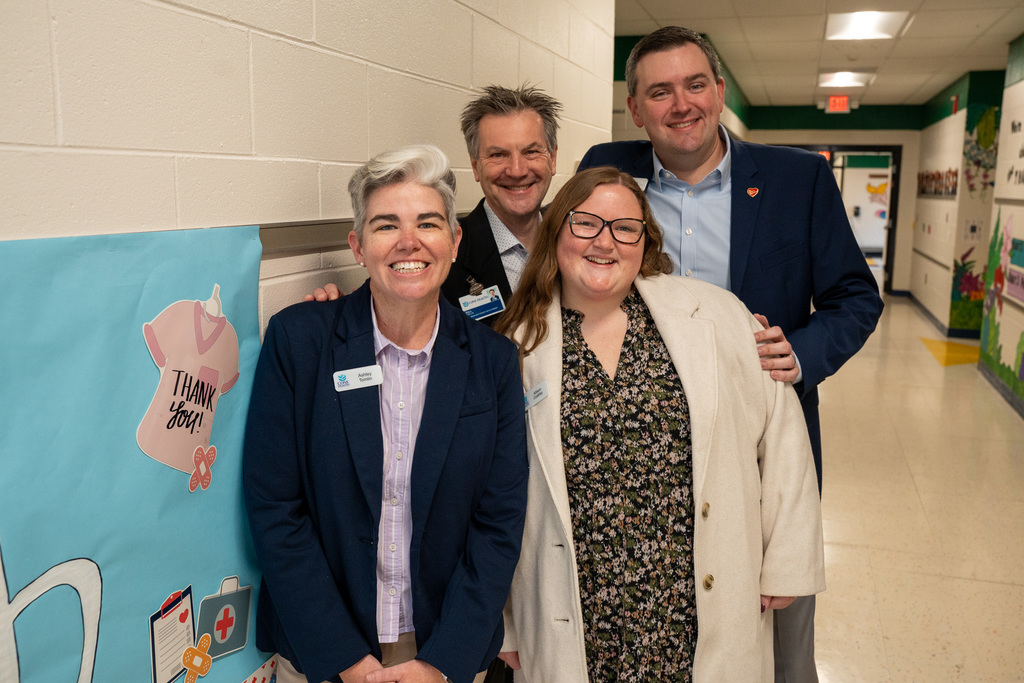 Group photo in a school hallway showing four smiling adults: two women in the front (one with short gray hair, one in a floral top and light jacket) and two men slightly behind them, all likely Cone Health and school partners at the event.