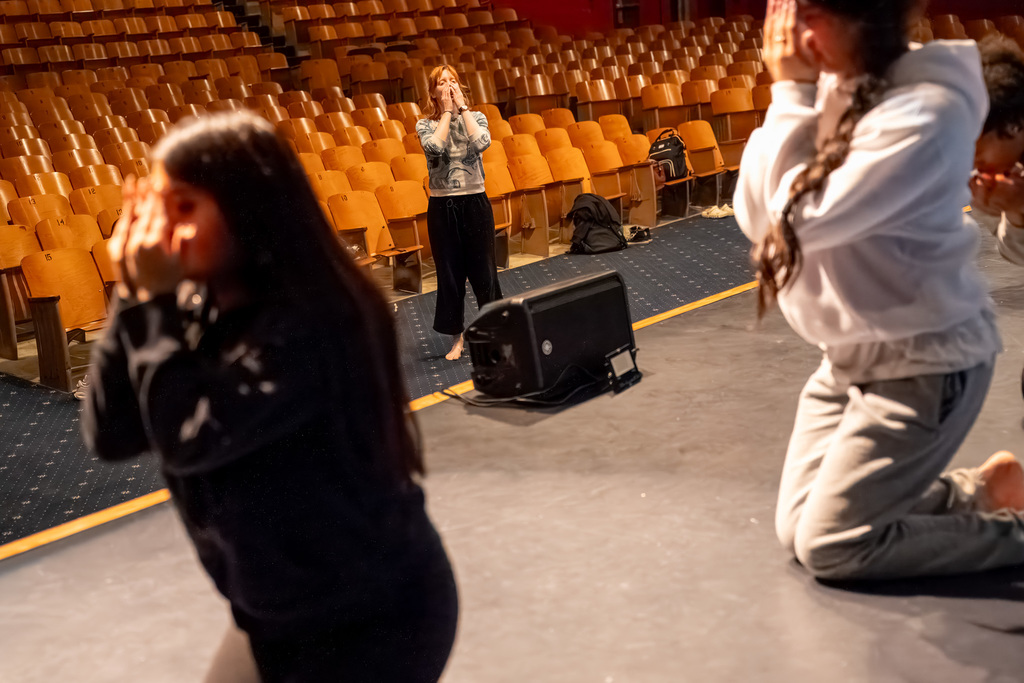 Dance students in a large auditorium practice a routine on the stage, kneeling with hands near their faces, while their instructor (Ms. Teachout) watches from the middle of the stage. This relates to preparing for the Winter Concert.