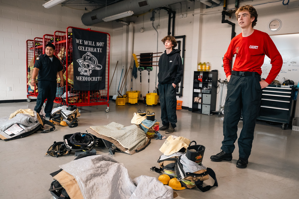 Two male students in Fire Academy casual uniform stand in a readiness exercise room with multiple piles of disassembled protective gear (turnout gear) on the floor, ready for them to practice dressing quickly.