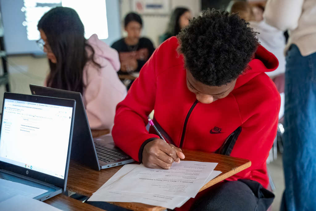 A male student in a red sweatshirt is leaning forward, intensely writing on a document at his desk with a pencil, a laptop open next to him. This represents a student hard at work in social studies or another class.