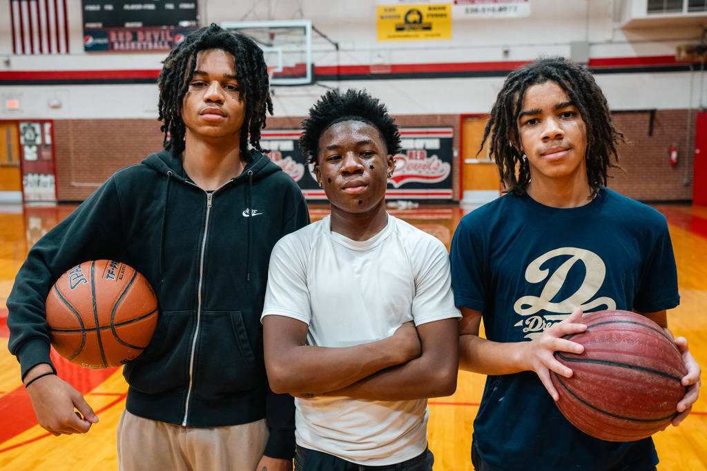 Three male students holding basketballs pose together on a wooden gymnasium floor. This relates to students working on their jumpers in PE with Ms. Egli.