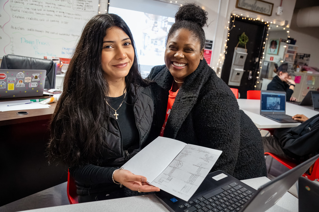 A smiling female student and a teacher (Ms. Surgeon) pose together in a brightly lit classroom, holding up a drawing of a floor plan with notes on it, related to interior design and living costs research.