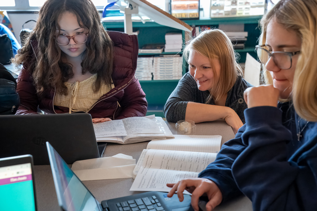 A teacher (likely Ms. Walsh or Ms. Daniels) is seated at a table, smiling at two female students who are looking down at open notebooks and working on laptops. This relates to the AP Government Blooket review or Ms. Daniels' social studies class.