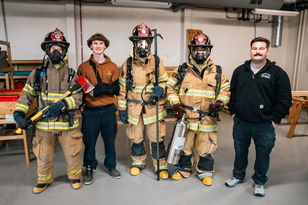 Three students wearing full Fire Academy protective gear, including masks and helmets, stand with firefighting tools and instructor. This relates to the Fire Academy juniors' preparation time.