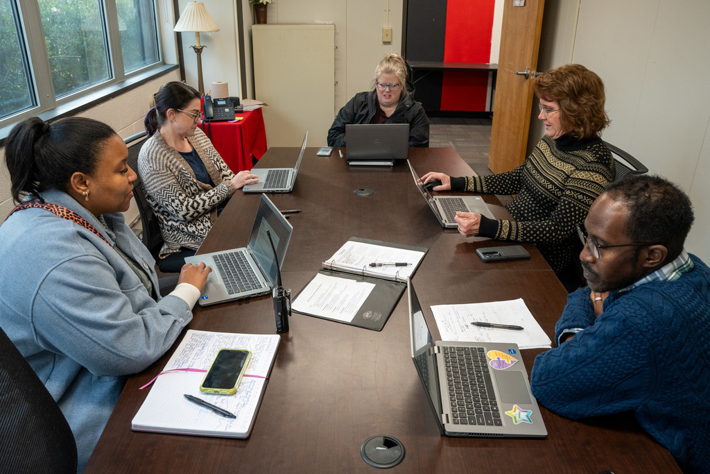 A group of six adults (three women and three men) are seated around a large conference table, all looking at their open laptops and discussing notes. This represents the Student Services team planning together.