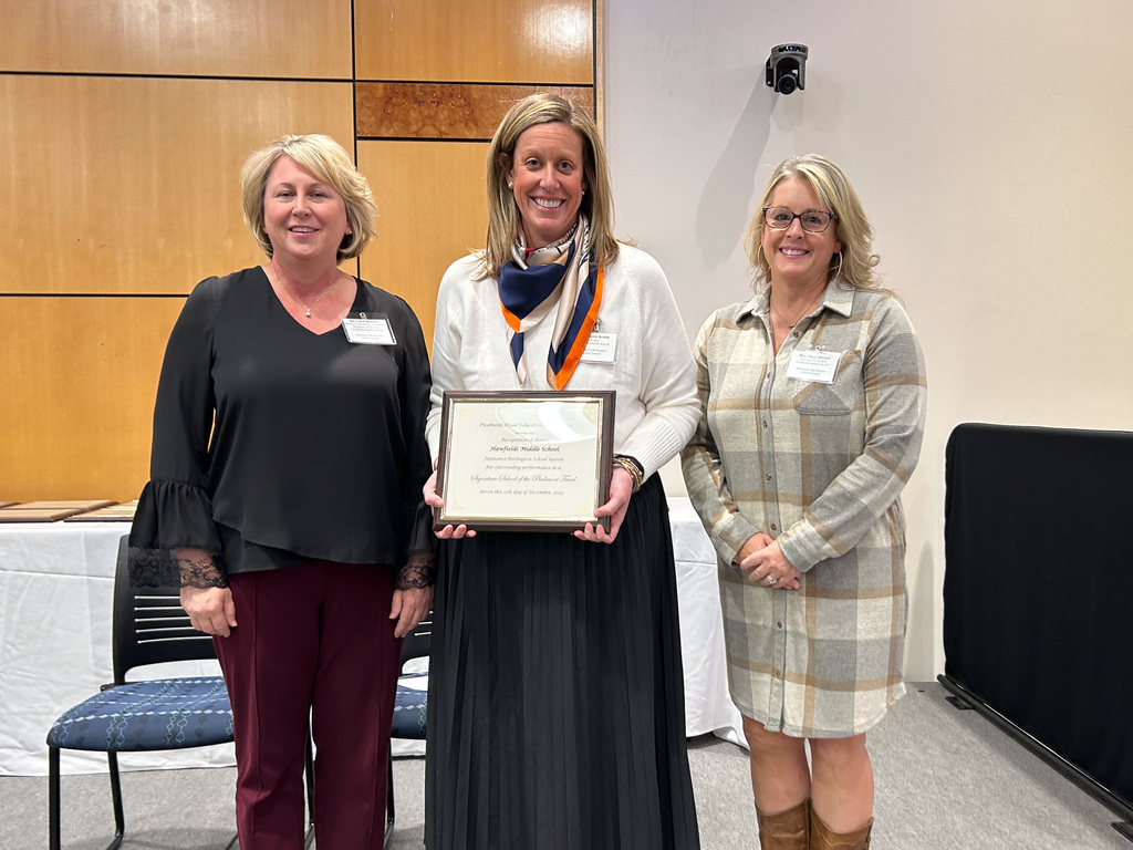 Three women stand together, with the woman in the middle holding a framed certificate. They are smiling at the camera after an award presentation.
