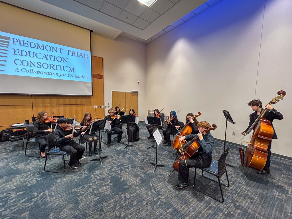 A student ensemble, primarily string players (violins, cellos, and a bass), performs on a blue carpeted stage area. A large screen in the background displays the text: "PIEDMONT TRIAD EDUCATION CONSORTIUM, A Collaboration for Education."