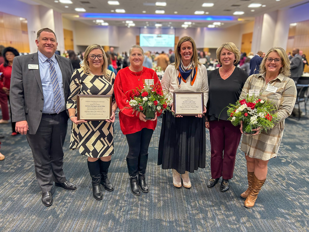 Six people—three men and three women—are standing side-by-side and smiling at the camera after a presentation. Two of the women hold floral arrangements, and two people are holding framed certificates. The event room is decorated for a holiday celebration.