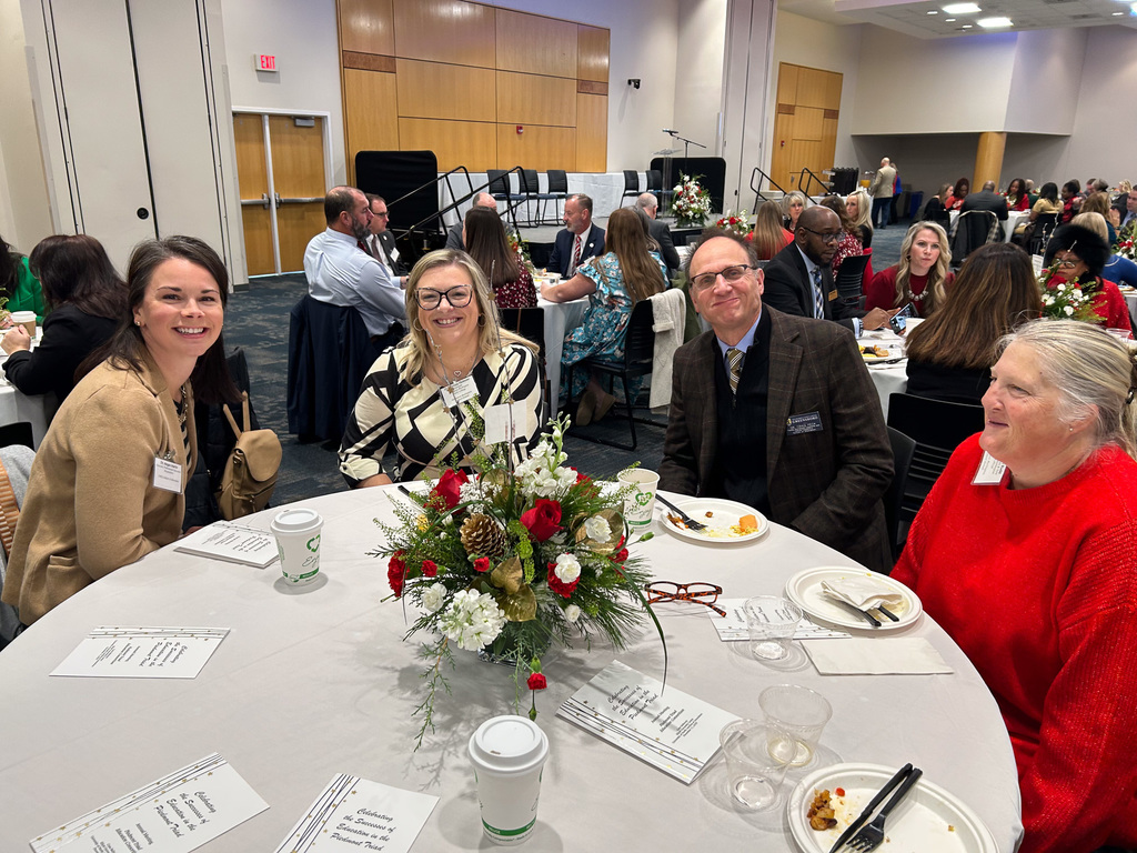 A group of five people, including two women smiling at the camera, are seated at a round table during a formal indoor event. A centerpiece of red and white flowers and greenery is in the middle of the table, which is set with coffee cups and printed programs.