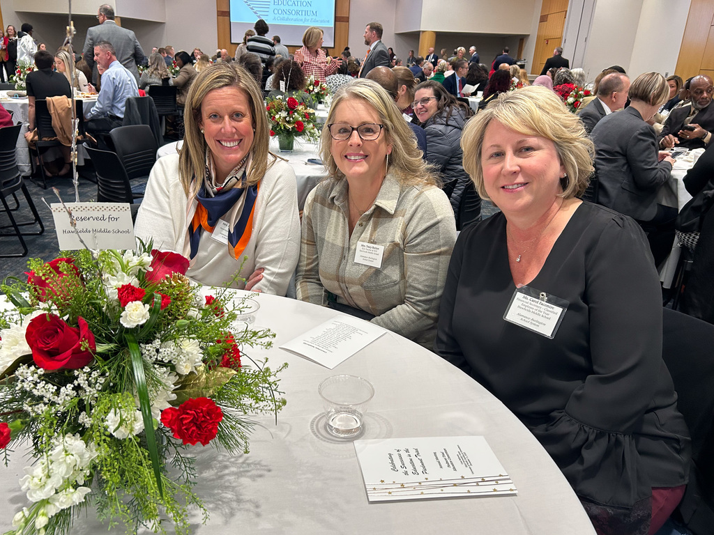 Three women are seated together at a round table reserved for "Hawfields Middle School." They are smiling at the camera, with a large, festive centerpiece of red and white flowers on the table in front of them. Other attendees are visible in the background.